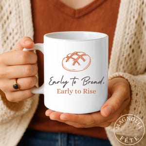 May include: White ceramic mug featuring a bread illustration and the text "Early to Bread. Early to Rise" in black and orange. The mug has a curved handle and is held by a person.