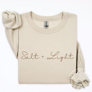 May include: A beige crew neck sweatshirt with the words "Salt + Light" in a brown script font. The sweatshirt is folded with the sleeves draped over the front. The brand name "Magnolia Fête" is visible in the lower right corner.