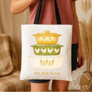 May include: White tote bag featuring a stack of three vintage-style casserole dishes in yellow, green, and cream. The top dish is yellow with white floral accents, the middle is green with chicken designs, and the bottom has wheat stalks. Text reads "Hot Dish Society."