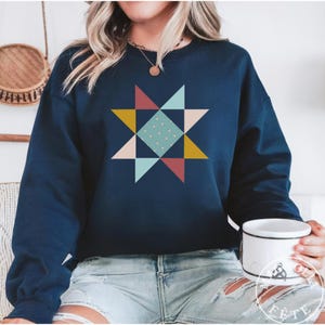 May include: Navy blue sweatshirt featuring a colorful geometric star design. The star is composed of triangles in shades of red, yellow, pink, and teal, with a dotted square in the center. The sweatshirt is a crew neck style.