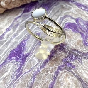 May include: A silver ring featuring a white, oval gemstone and a feather motif. The ring is displayed on a clear plastic stand, set against a purple and white marbled backdrop. The feather design adds a touch of nature and elegance to the jewellery.