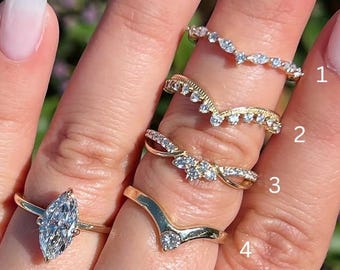 May include: A hand displays a collection of gold rings with various diamond and gemstone settings. The rings include a marquise-cut diamond solitaire, delicate bands with small stones, and a chevron-shaped ring. The background is a blurred view of green foliage.