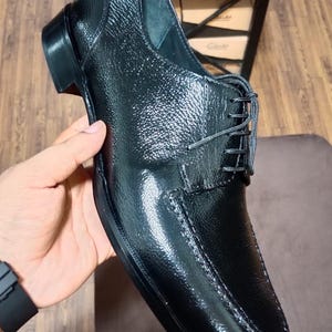 Pode incluir: Um sapato social de couro preto com acabamento texturizado e granulado e brilho intenso. O sapato apresenta um fecho de atacadores pretos finos, um design de biqueira costurada e um salto baixo. Um segundo sapato com sola vermelha é visível.