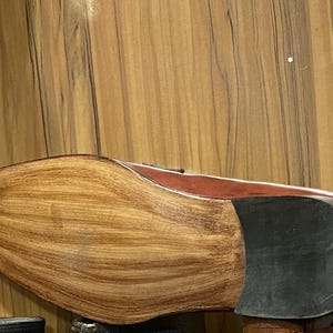 May include: A brown leather shoe featuring a polished, light-colored wooden sole with a distinct grain pattern and a solid black rubber heel cap. The shoe has a smooth, rounded toe profile and a sleek, structured design with visible stitching.