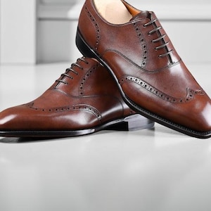 May include: A pair of brown leather dress shoes with intricate brogue detailing and a classic lace-up design. The shoes are displayed on a white surface, showcasing their polished finish and elegant style.