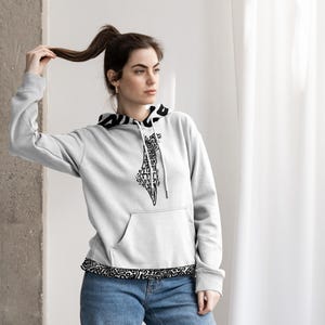 May include: A pale grey hoodie with a black and white patterned trim and a black graphic design on the front. The hoodie has a front pocket and a drawstring. The person in the image is wearing blue jeans.