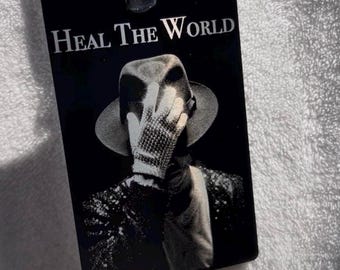 May include: A rectangular black badge featuring a white graphic of a figure wearing a fedora and a sequined glove covering their face. The text HEAL THE WORLD is printed in white at the top. A black metal clip is attached to the top edge of the badge.