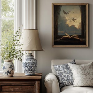 May include: A cozy living room scene featuring a framed artwork with dragons flying in a cloudy sky. A blue and white patterned lamp sits on a wooden nightstand, next to a vase with greenery. A white sofa with patterned pillows completes the scene.