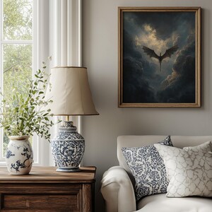 May include: A framed artwork of a dragon in a cloudy sky hangs above a white sofa with patterned cushions. A wooden bedside table supports a blue and white patterned lamp and vase. The room has a neutral colour palette.