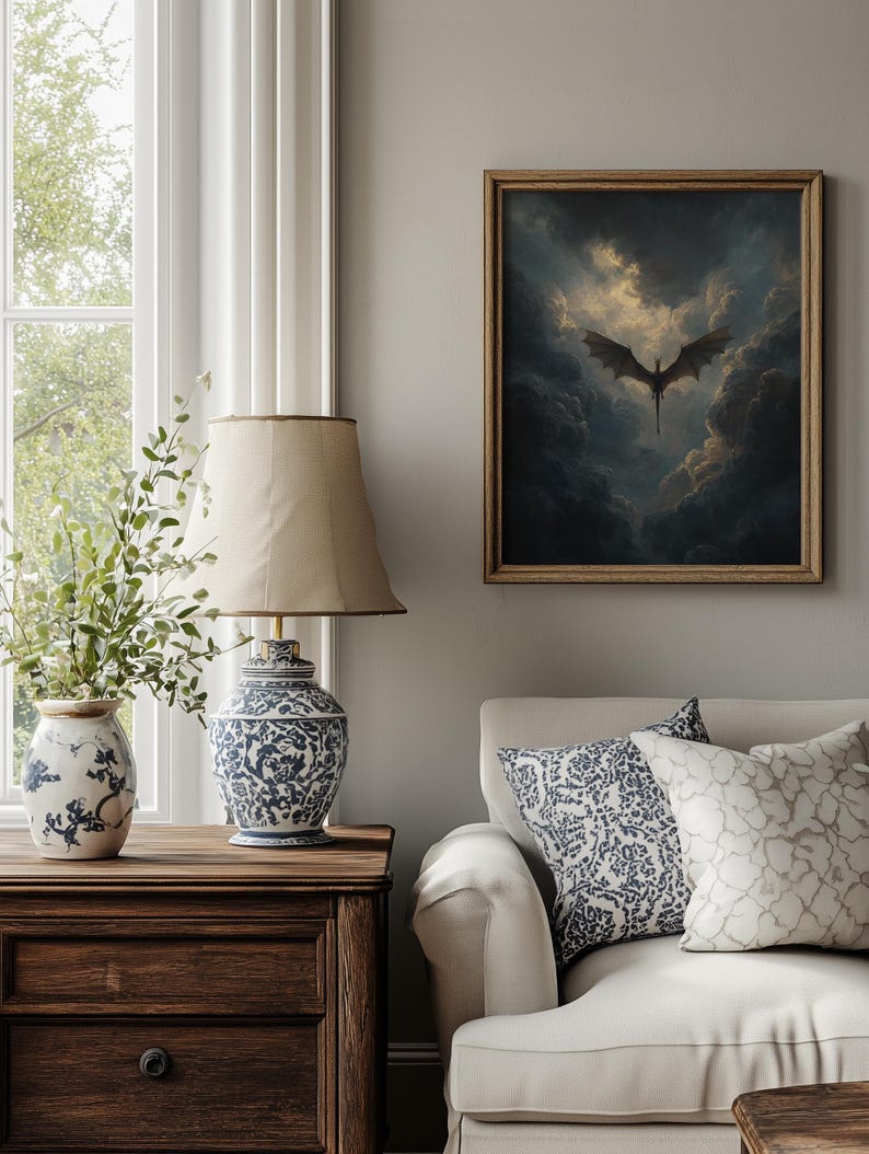 May include: A framed artwork of a dragon in a cloudy sky hangs above a white sofa with patterned cushions. A wooden bedside table supports a blue and white patterned lamp and vase. The room has a neutral colour palette.