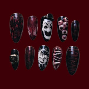 Puede incluir: Un conjunto de diez uñas postizas largas y ovaladas sobre un fondo rojo oscuro. Las uñas presentan varios diseños, incluyendo una cara de payaso, globos oculares y texto. Algunas uñas tienen un efecto de sangre, mientras que otras tienen un aspecto texturizado.
