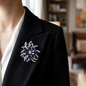 Peut inclure: Une broche florale avec un centre bleu foncé et des accents de cristaux transparents. La broche est épinglée sur un blazer noir, porté sur une chemisier blanc. La broche a un sertissage en métal argenté.