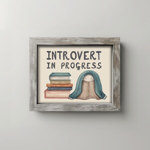 May include: A framed print with the text "INTROVERT IN PROGRESS." The artwork features a stack of books and a cartoon figure wrapped in a blue blanket. The frame is made of weathered gray wood.
