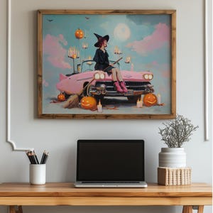 May include: A framed artwork depicts a witch in a black dress and hat, seated on a pink vintage car. The Halloween-themed scene includes jack-o'-lanterns, candles, and a full moon in a pastel sky. A broom rests on the car.