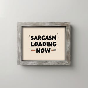 May include: A framed print with the text "SARCASM LOADING - NOW -" in black, with orange accents. The print is set against a cream background and framed in a weathered gray wood.