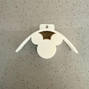 May include: A white, 3D-printed wall-mounted holder featuring a Mickey Mouse head design. The holder has a circular center with two rounded ears and two curved wings extending outward. It includes a small hole for hanging.