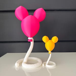 May include: Two decorative sculptures shaped like balloons with rounded ears. The larger sculpture is bright pink, and the smaller one is yellow. Both balloons are attached to white, coiled rope-like bases that spiral outward at the bottom.