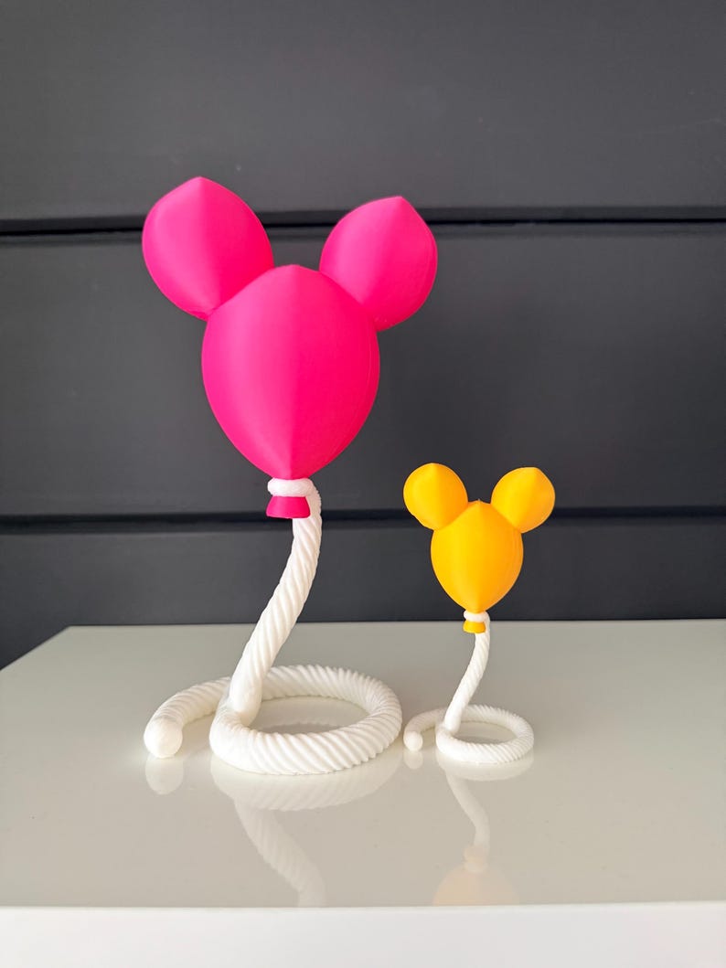 May include: Two decorative sculptures shaped like balloons with rounded ears. The larger sculpture is bright pink, and the smaller one is yellow. Both balloons are attached to white, coiled rope-like bases that spiral outward at the bottom.