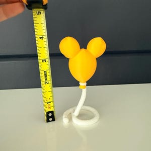 May include: A yellow balloon-shaped object with two rounded ears at the top, attached to a white coiled rope base. A yellow measuring tape is positioned next to the object, showing a total height of approximately 4.5 inches from base to top.