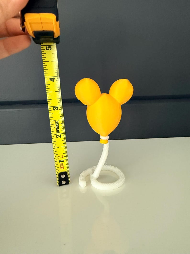 May include: A yellow balloon-shaped object with two rounded ears at the top, attached to a white coiled rope base. A yellow measuring tape is positioned next to the object, showing a total height of approximately 4.5 inches from base to top.
