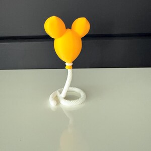 May include: A bright yellow balloon-shaped object with two rounded ears at the top, resembling a mouse head silhouette. The balloon is attached to a white, coiled, rope-like base that curves along the surface, providing a stable stand.