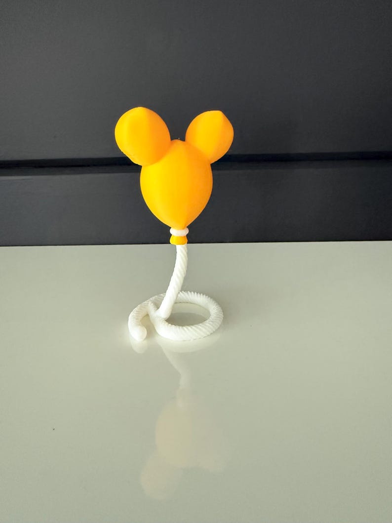 May include: A bright yellow balloon-shaped object with two rounded ears at the top, resembling a mouse head silhouette. The balloon is attached to a white, coiled, rope-like base that curves along the surface, providing a stable stand.
