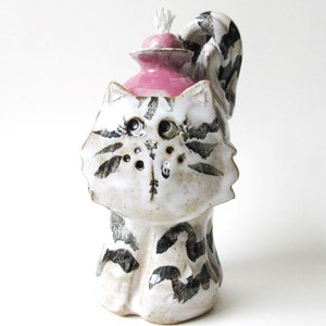 May include: A white ceramic cat figurine with black markings, holding a pink oil lamp with a white wick. The cat has a whimsical expression with black eyes and a small, black nose.
