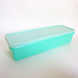 May include: A rectangular, turquoise plastic container with a translucent lid. The container is long and narrow, with a slightly rounded edge. The lid fits snugly on top, creating a seal. The container is empty and sits on a white surface.