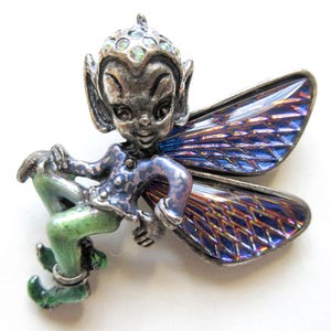 May include: A silver-toned fairy brooch with a whimsical design. The fairy has a blue jacket, green pants, and iridescent blue wings. The fairy's head is adorned with small, sparkling accents. A charming accessory for a jacket or bag.
