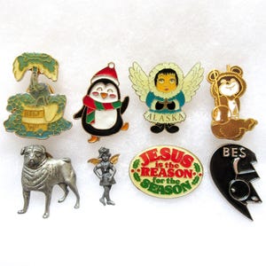 May include: A collection of enamel pins featuring various designs. Includes a penguin in a Santa hat, a pug, an angel, a bear, and a pin with the text "JESUS is the REASON for the SEASON".