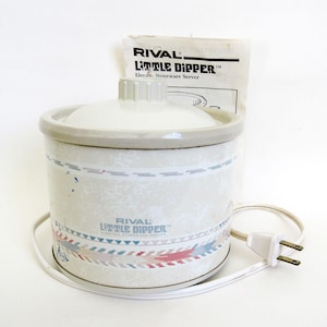 May include: A white Rival Little Dipper electric stoneware server with a lid and a decorative band of blue, red, and gray. The appliance has a white power cord and a two-prong plug. The product's name is printed on the side.