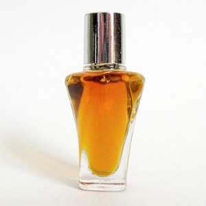 May include: A small, clear glass perfume bottle with a tapered, angular body filled with amber-colored liquid. The bottle is topped with a cylindrical, reflective silver-toned metal cap that sits flush against the neck of the glass container.