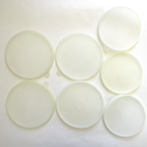 May include: A collection of seven off-white, circular plastic lids. Each lid has a slightly raised edge and a small tab for easy removal. The lids are arranged on a white surface, providing a clean and simple aesthetic.
