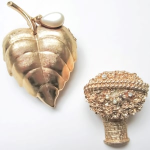 May include: Two gold tone brooches. One is a leaf with a pearl accent. The other is a basket of flowers with rhinestones.