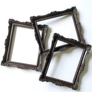 May include: Three rectangular picture frames with a dark brown finish and ornate, raised floral and scrollwork detailing along the edges. The frames are arranged in an overlapping cluster, each featuring a hollow center and a decorative border.