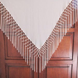 May include: A triangular off-white fabric shawl featuring an intricate macrame border along the two angled edges. The border consists of a knotted net pattern that transitions into long, straight, hanging fringe tassels in a matching off-white color.