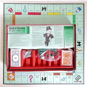 May include: A Monopoly board game set, featuring a green game board with property spaces and a red tray containing game pieces, cards, and dice. The box includes text about the game rules and facts.