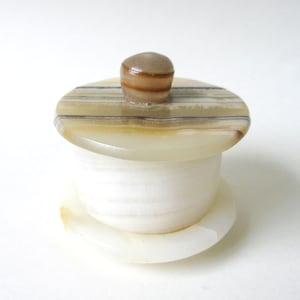 May include: A small, round, white alabaster trinket box with a striped lid and a brown knob. The box is made of white alabaster with brown stripes.
