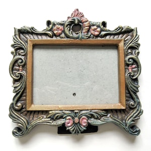 May include: Ornate, rectangular picture frame with a gold-colored inner frame and a decorative outer frame in shades of green, bronze, and pink. The frame features floral and scrollwork details, and a black stand.