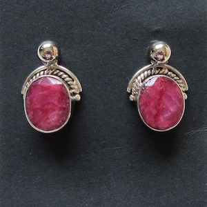 May include: A pair of silver earrings with a round, pink ruby gemstone set in each. The earrings have a simple, elegant design with a small silver ball at the top.