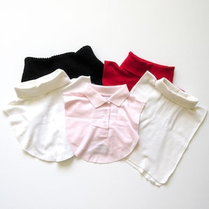 May include: A collection of detachable shirt collars in various colors. The collars include a black ribbed turtleneck, a red turtleneck, and white and pink collared shirts with button details. The collars are arranged on a white surface.
