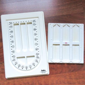 May include: Two off-white plastic craft tools. One is a rectangular tray with a curved bottom, numbered markings, and three long slots. The other is a smaller, square tray with six rectangular compartments. The brand name "Plagfime" is visible.