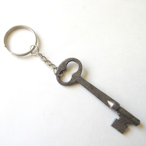 May include: A vintage-style key attached to a silver key ring and chain. The key is made of a dark, aged metal with a decorative, openwork design at the top. The key's rectangular bit is visible at the bottom.