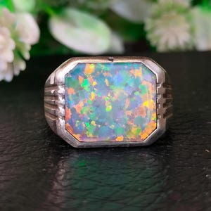 Op de afbeelding: Een zilveren ring met een grote, rechthoekige opaalsteen. De opaal toont een levendig kleurenspel, waaronder blauwe, groene, oranje en gele tinten. De ring heeft een eenvoudig, maar elegant ontwerp, geschikt voor dagelijks gebruik.