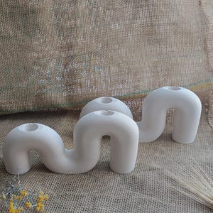 May include: Three white ceramic candle holders with a unique, curved design. The holders have a smooth, matte finish and a modern, minimalist aesthetic. They are arranged on a textured, neutral-toned surface, with a few sprigs of dried flowers.