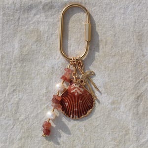 Full length flatlay of strawberry quartz chip bead and freshwater pearl strand alongside natural scallop shell pendant with gold bow charm hanging from gold oval carabiner clasp on linen