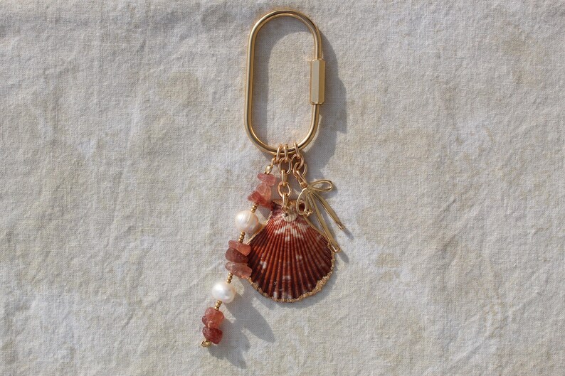 Full length flatlay of strawberry quartz chip bead and freshwater pearl strand alongside natural scallop shell pendant with gold bow charm hanging from gold oval carabiner clasp on linen
