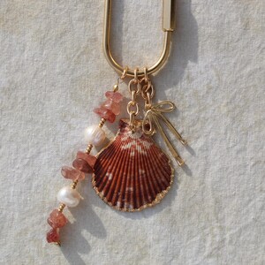 Close up of strawberry quartz chip beads alternating with freshwater pearls alongside large natural scallop shell with gold dipped edges and gold bow charm on linen