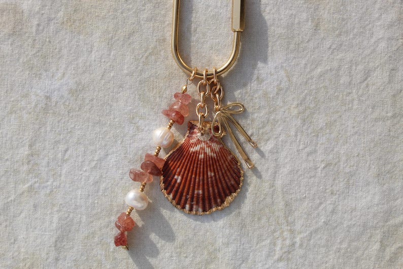 Close up of strawberry quartz chip beads alternating with freshwater pearls alongside large natural scallop shell with gold dipped edges and gold bow charm on linen