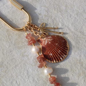 Angled view of full coastal bag charm keychain showing gold carabiner clasp, strawberry quartz chip beads, freshwater pearls and natural scallop shell pendant on linen
